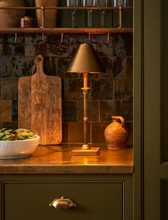 Kelmscot table lamp in antique brass lit on a kitchen worktop in a green painted kitchen