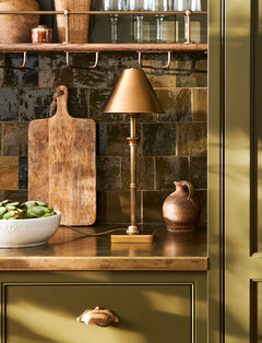 Antique brass Kelmscot table lamp lit on a wooden kitchen worktop beside a chopping board