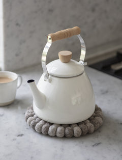 White enamel stove kettle resting on a felt trivet on a marble worktop beside a mug of tea